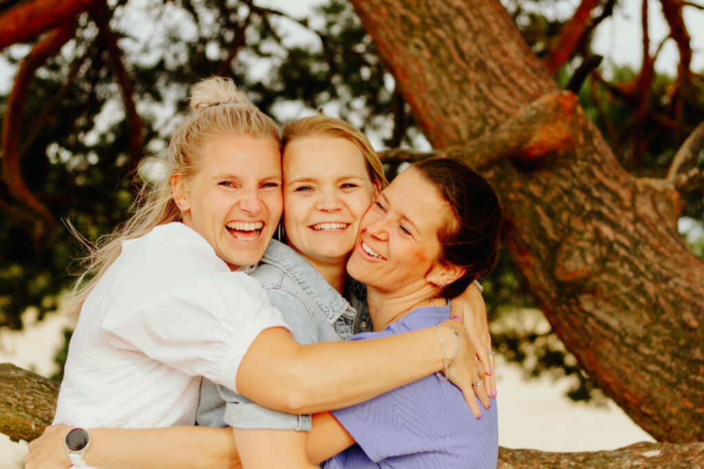 drie vrouwen knuffelend op de foto voor een boom op een zanderstuiving tijdens een fotoshoot.